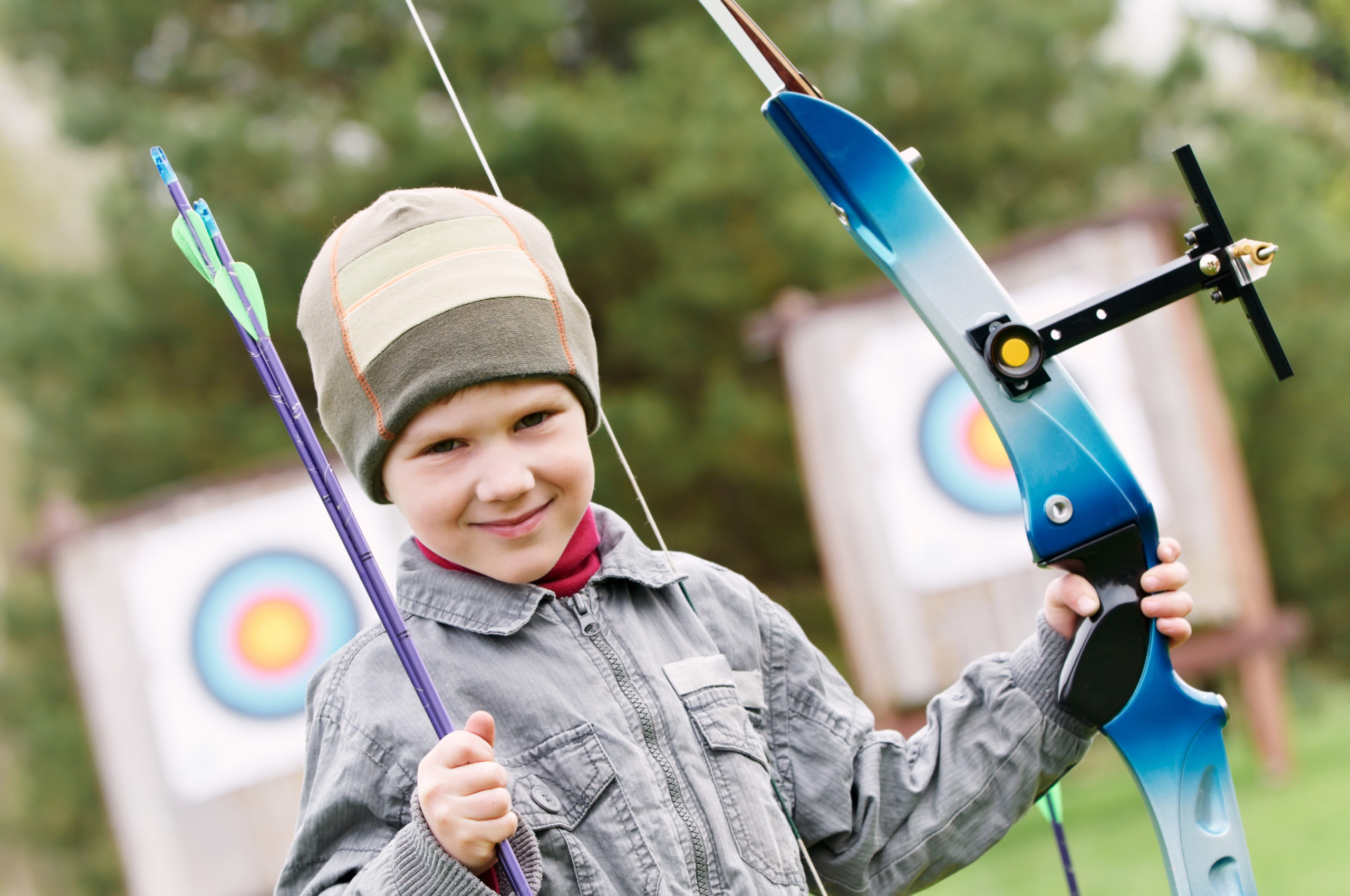 Smiling child archer with bow and arrows in front of target spring outdoors