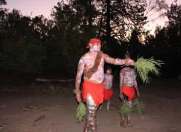 Men covered in white paint wearing red head piece and red cloth holding leaves